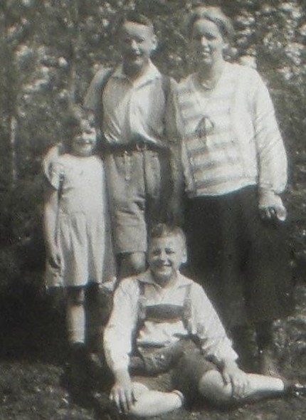 Picture of Marie and her children. She is standing to the right. Her son Karl next to her in short pants. He has wrapped his arm around his younger sister. The younger son Helmut sits on the ground wearing what looks like Lederhosen. Everybody is smiling. It appears to be a warm day. They are out in the garden or a park. 